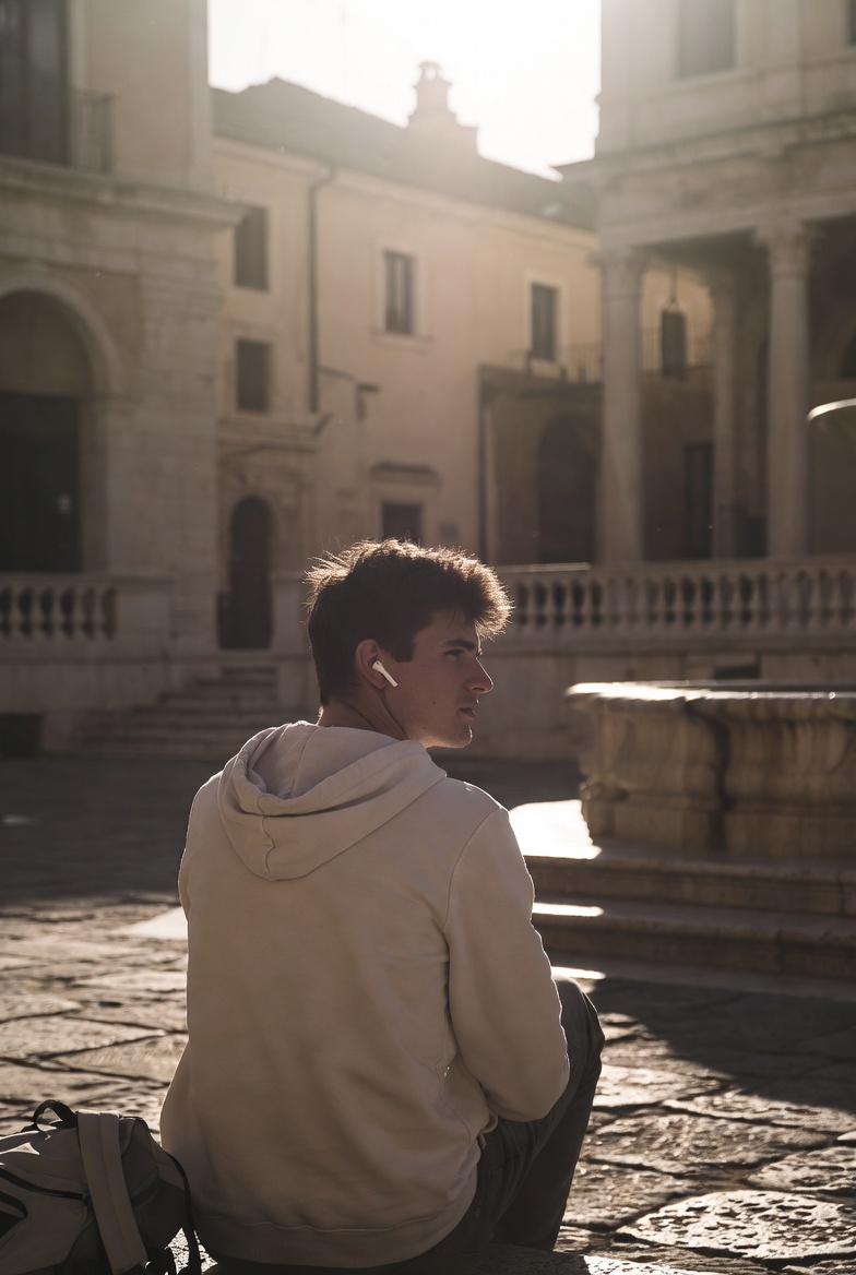 A young man sitting alone in a sunlit Italian piazza, unhurried, listening with an earbud